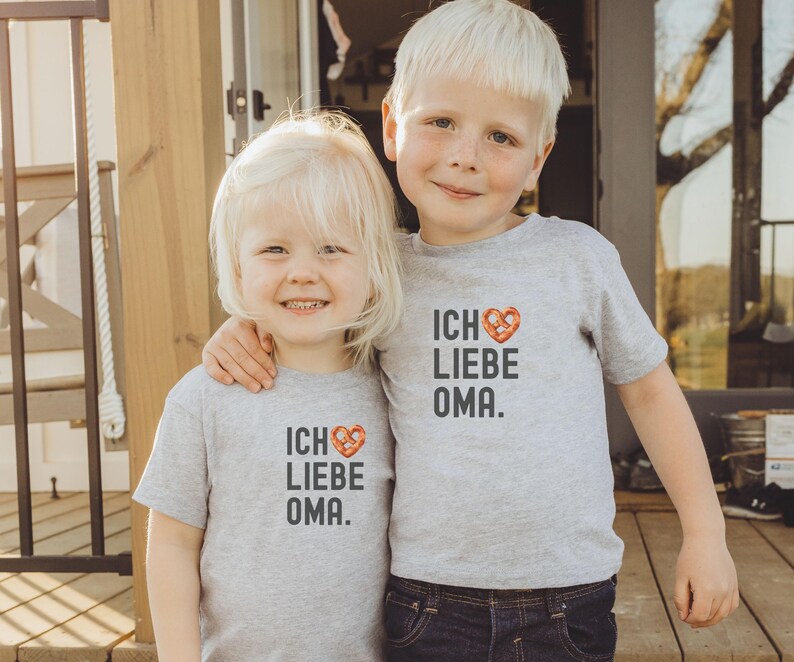 May include: Two children wearing grey t-shirts with the text "Ich Liebe Oma" and a pretzel heart graphic. The child on the left is a girl and the child on the right is a boy.