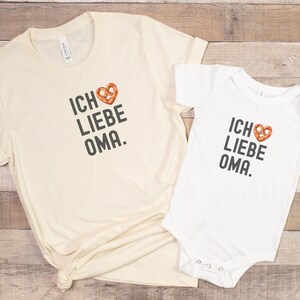 May include: Two white t-shirts with the text "Ich liebe Oma" and a pretzel heart graphic. The larger shirt is a long-sleeved t-shirt and the smaller shirt is a short-sleeved bodysuit.