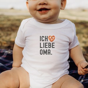 May include: A white baby onesie with the text "ICH LIEBE OMA." in black lettering. A pretzel in the shape of a heart is above the word "LIEBE".
