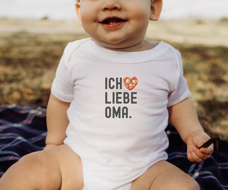 May include: A white baby onesie with the text "ICH LIEBE OMA." in black lettering. A pretzel in the shape of a heart is above the word "LIEBE".