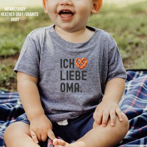 May include: A toddler wearing a dark heather grey t-shirt with the text "Ich liebe Oma." and a pretzel heart graphic. The toddler is sitting on a plaid blanket.