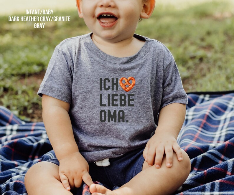 May include: A toddler wearing a dark heather grey t-shirt with the text "Ich liebe Oma." and a pretzel heart graphic. The toddler is sitting on a plaid blanket.