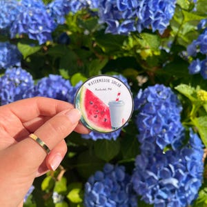 May include: A round sticker featuring a watercolor illustration of a watermelon slice and a drink with a red and white striped straw. The text "Watermelon Sugar" and location are printed on the sticker. The sticker is held in front of a backdrop of blue hydrangea flowers.