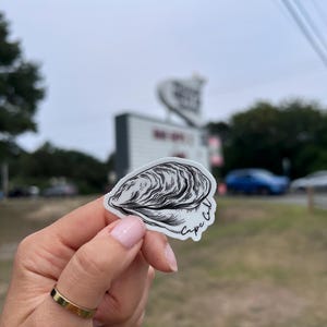 May include: A white sticker featuring a detailed black and white illustration of an oyster shell. The sticker has the words "Cape Cod" in a handwritten font. The sticker is held up in front of a blurred outdoor background.