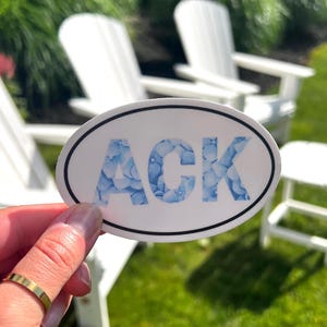 May include: An oval-shaped sticker with a white background and a thin black border. The letters ACK are printed in the center, filled with a pattern of light blue hydrangea flowers. The sticker is held against a background of white outdoor chairs.