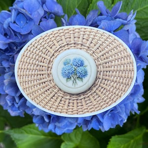 May include: An oval-shaped brooch featuring a woven tan wicker-style texture with a white circular center inlay. The inlay displays a detailed illustration of a cluster of blue hydrangea flowers with green stems and leaves on a white background.