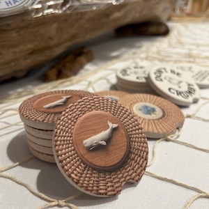 Pode incluir: Porta-copos redondos de cer&acirc;mica para carro com um c&iacute;rculo central de gr&atilde;o de madeira e uma figura de baleia branca em relevo, rodeados por uma borda bege texturizada com efeito de vime. Alguns exibem um design floral azul ou o texto Don't Be Crumbly.
