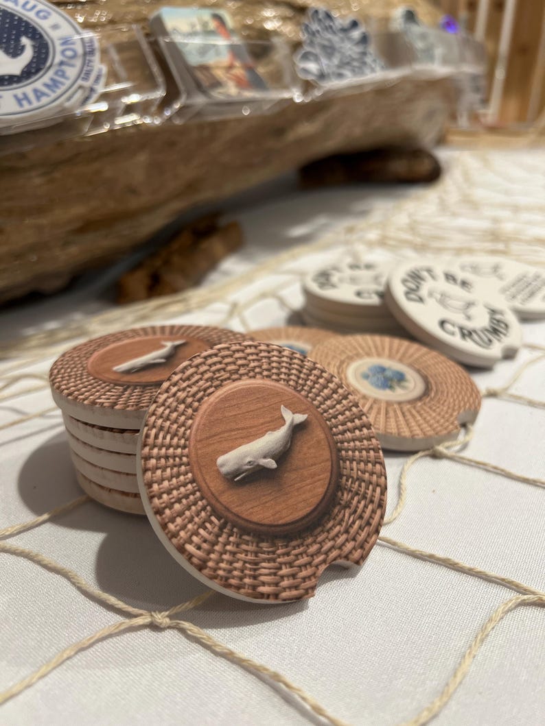 Pode incluir: Porta-copos redondos de cer&acirc;mica para carro com um c&iacute;rculo central de gr&atilde;o de madeira e uma figura de baleia branca em relevo, rodeados por uma borda bege texturizada com efeito de vime. Alguns exibem um design floral azul ou o texto Don't Be Crumbly.