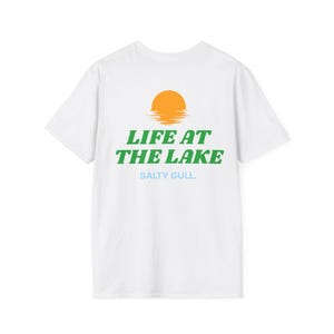 May include: A white short-sleeve t-shirt featuring a graphic on the back. The design includes an orange sun setting over water, the green text LIFE AT THE LAKE, and the light blue text SALTY GULL. The shirt has a crew neckline and short sleeves.
