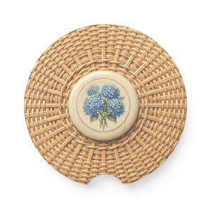 Puede incluir: Posavasos redondo con diseño de ratán tejido. El centro presenta un inserto circular de cerámica blanquecina con una ilustración floral de hortensias azules. El posavasos tiene una pequeña muesca en un lado.