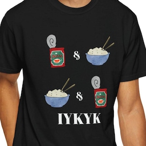 以下が含まれることがあります： 黒いTシャツ。豆の缶と箸の付いたご飯のボウルが描かれたグラフィックデザイン。テキスト「IYKYK」がTシャツの下部に白で印刷されています。