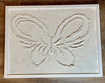 May include: A white framed canvas with a textured, raised butterfly design in the center. The butterfly is outlined in white and has a textured, raised body.