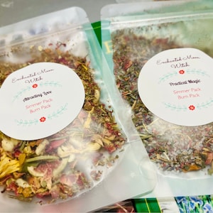 May include: Two clear plastic bags filled with dried herbs and flowers. The bags are labeled "Enchanted Moon Witch" and have different titles: "Attracting Love" and "Practical Magic". Both bags have the text "Simmer Pack Burn Pack".
