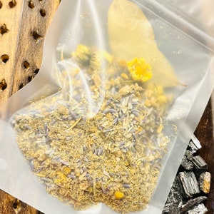May include: A clear plastic bag filled with dried herbs and flowers, including lavender, chamomile, and bay leaves. The bag is sitting on a wooden surface next to a bowl of black charcoal.