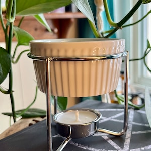 May include: A white ceramic wax warmer with a ribbed design, held by a silver metal stand. A lit tea light candle sits below. The warmer is surrounded by green leafy plants.