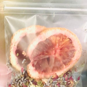 May include: A clear plastic bag filled with dried herbs and flowers, including lavender, rose petals, and other botanicals. Two halves of a blood orange are on top of the mixture.