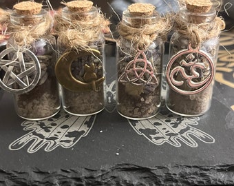 May include: Four small glass bottles with cork tops and twine accents. Each bottle contains a different charm: a pentagram, a crescent moon, a triquetra, and an om symbol. The bottles are arranged on a black slate surface with a butterfly design.