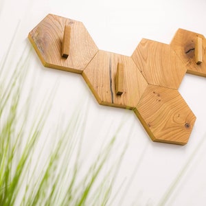 May include: Wooden wall-mounted coat rack in a honeycomb design. The rack is made of light-coloured wood and features six hexagonal pieces with wooden pegs for hanging items. The rack is mounted on a white wall.