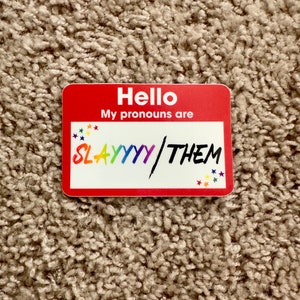 May include: Red name tag with the text "Hello, My pronouns are SLAYYYY/THEM" in rainbow colors with stars around the text.