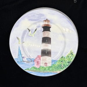 May include: A set of decorative plates featuring a lighthouse scene. The plates depict a black and white striped lighthouse, a pink building, sailboats, seagulls, and a blue ocean under a cloudy sky. The plates have a clear, glossy finish.