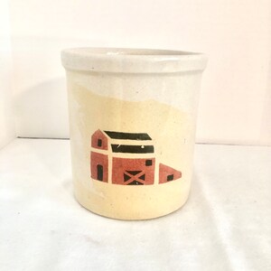 May include: A white ceramic utensil holder with a red barn design. The barn has a black roof and a white window.