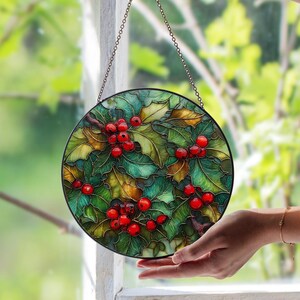 May include: Circular stained glass panel depicting holly with green leaves and red berries. The panel is framed in black and suspended by a chain. The background shows blurred green foliage.