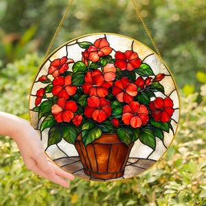 May include: A circular stained glass suncatcher featuring a vibrant arrangement of red flowers with green leaves in a brown pot. The piece is framed in a gold-coloured metal border and includes a gold chain for hanging, set against a clear background.