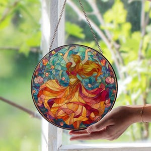 May include: Round stained glass window art featuring a dancing woman with flowing red and yellow hair and dress. The artwork is in a mosaic style with a black metal frame and hanging chain. The background is a mix of blue, green, and orange.