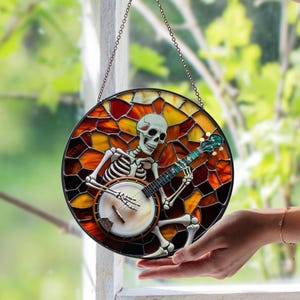 May include: Round stained glass window art featuring a skeleton playing a banjo. The skeleton is white, with a banjo in its hands. The background is a mosaic of orange, yellow, and red glass pieces, with a black border. The window hangs from a chain.