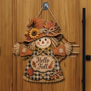 Könnte beinhalten: Ein dekorativer Vogelscheuchen-Wandschmuck mit einem braunen Hut, der mit einer Sonnenblume und Ahornblättern verziert ist, einem karierten Kleid mit einem Juteschild mit der Aufschrift Hello Fall und strohartigen Fransen an Ärmeln und Saum.