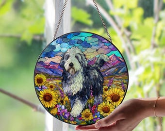 May include: Round stained glass window art featuring a dog in a field of sunflowers and wildflowers, with mountains in the background. The dog has black and white fur. The window is hanging from a chain.