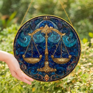 May include: Round stained glass Libra zodiac sign with a gold-colored frame and hanging chain. The artwork features a gold balance scale against a blue and teal background with a starry night design. The image is held in front of a green, leafy background.