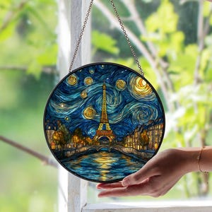 May include: Round stained glass window art depicting the Eiffel Tower in Paris under a swirling night sky, reminiscent of Van Gogh's style. The artwork features shades of blue, yellow, and orange, with a black metal frame and hanging chain.