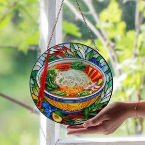 May include: Round stained glass window depicting a bowl of ramen with red chopsticks. The bowl is filled with noodles, vegetables, and broth. The window is framed with colorful glass pieces and hangs from a chain.