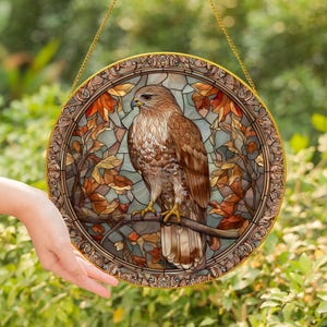 May include: Circular stained glass window depicting a hawk on a branch. The artwork has a detailed, mosaic-like design with brown, orange, and green hues. A gold chain is attached for hanging. The background is a blurred green.