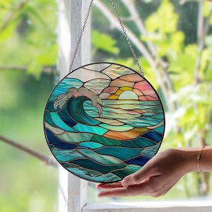 May include: Round stained glass panel depicting a seascape with a breaking wave and a sunset. The artwork features shades of blue, green, orange, and pink, with a black metal frame and a chain for hanging. The panel is held by a hand.