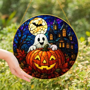 Può includere: Un acchiappasole circolare in vetro colorato raffigurante una zucca sorridente con un fantasma sopra. Lo sfondo mostra un cielo notturno blu scuro, una luna piena con la sagoma di un pipistrello, una casa stregata e foglie autunnali arancioni.