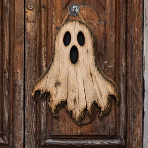 May include: A ghost-shaped wall hanging crafted from light-coloured wood with a visible grain pattern. The cutout features two oval eyes and an oval mouth, all backed by a dark material. The edges are darkened, and it hangs from a jute rope.