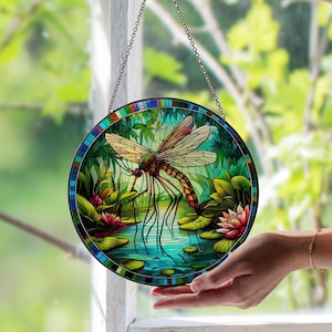 Op de afbeelding: Ronde gebrandschilderde glazen raamkunst met een libelle in een vijver. De libelle heeft bruine vleugels en een rood en groen lichaam. De vijver heeft waterlelies en roze bloemen. Het raam heeft een kleurrijke rand en hangt aan een ketting.
