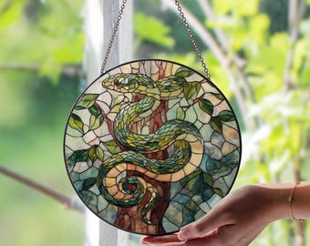 May include: Circular stained glass panel depicting a green snake coiled around a tree. The artwork is framed in black metal and hangs from a chain. The snake is rendered in shades of green and yellow, with a backdrop of leaves and a brown tree trunk. The panel is approximately 20 cm in diameter.