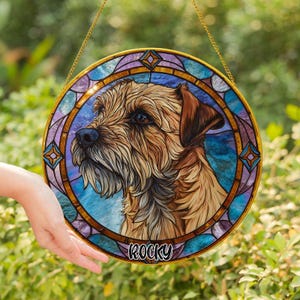 May include: A round stained glass window featuring a detailed portrait of a dog with brown and tan fur. The dog's name, "Rocky," is written in white script at the bottom. The window has a gold-colored border and is suspended by a gold chain.
