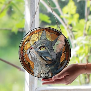 May include: Round stained glass window art featuring a cat with gray and blue fur, yellow eyes, and the name "BELLA" below. The cat is set against a warm, amber-toned background. A chain is attached for hanging.