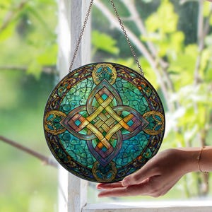 May include: Circular stained glass window with a Celtic knot design. The window features a central knot in gold and teal, set against a teal and green background. It hangs from a chain. The window is displayed in front of a window.