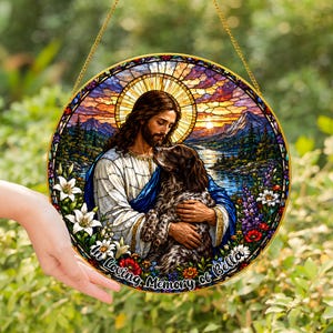 May include: A circular stained glass style suncatcher featuring a figure with long hair and a beard holding a brown dog. The scene includes a mountain lake landscape, white lilies, and the text In Loving Memory of Bella at the bottom.