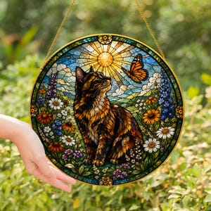 May include: Circular stained glass panel depicting a cat, butterfly, and floral scene. The cat has brown and orange markings, looking up at a butterfly. The background shows a sun, blue sky, and various flowers. A gold chain is attached for hanging.