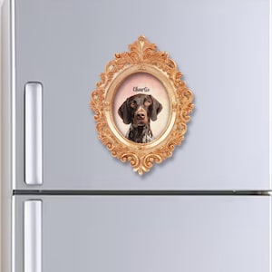 May include: A fridge magnet with a portrait of a German Shorthaired Pointer named Charlie, set in an ornate, gold-coloured oval frame. The dog has a brown coat with white markings. The frame is attached to a refrigerator.