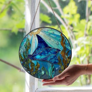 以下が含まれることがあります： 青いマンタが水中で泳ぐ様子を描いた丸いステンドグラスパネル。青、ティール、ゴールドの色合いで太陽光線、泡、海藻が描かれています。パネルは黒いフレームで縁取られ、チェーンで吊り下げられています。