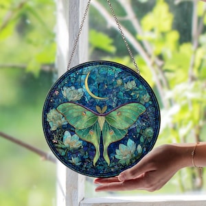May include: Round stained glass window art featuring a green moth with detailed wings, a crescent moon, and floral accents. The artwork is predominantly blue and green, with a chain for hanging. The design is reminiscent of a mosaic.