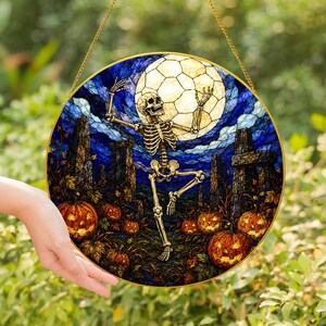 Può includere: Un acchiappasole circolare in vetro colorato raffigurante uno scheletro che danza davanti a una grande luna piena. La scena include nuvole blu scuro, pilastri di pietra e diverse zucche di Halloween arancioni luminose, il tutto incorniciato in oro.