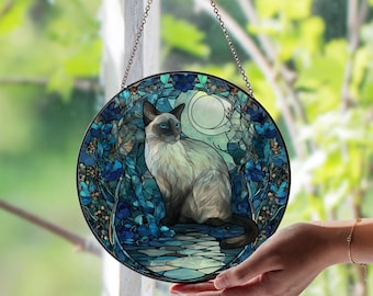 May include: Round stained glass window art featuring a Siamese cat in a moonlit garden scene. The artwork is predominantly blue and teal, with a detailed floral border and a chain for hanging. The cat has blue eyes and is sitting.